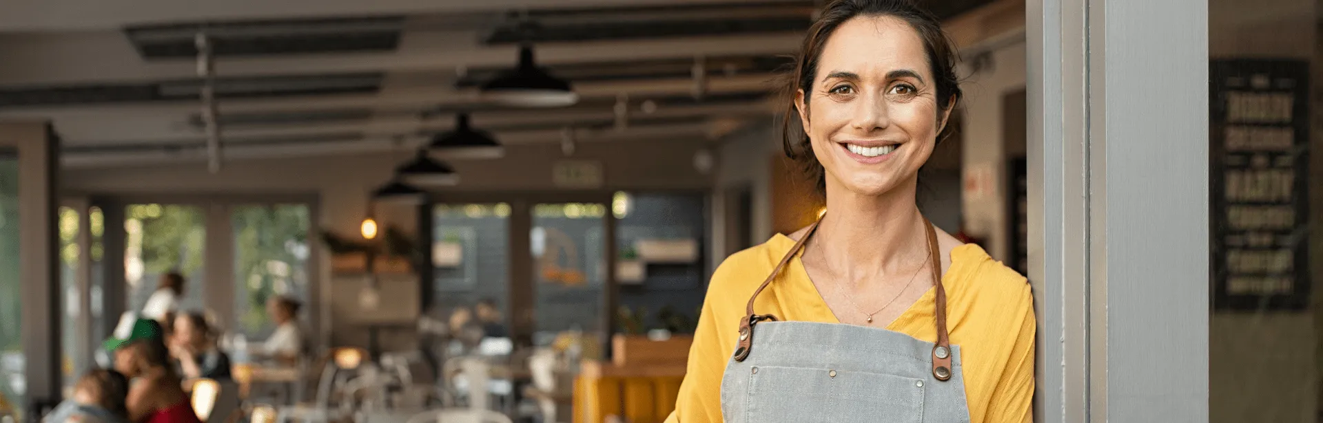 A woman wearing an apron in a coffee shop with customers in the background.