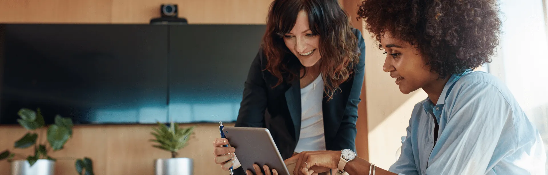 Two women in an office setting, smiling at a tablet computer screen.